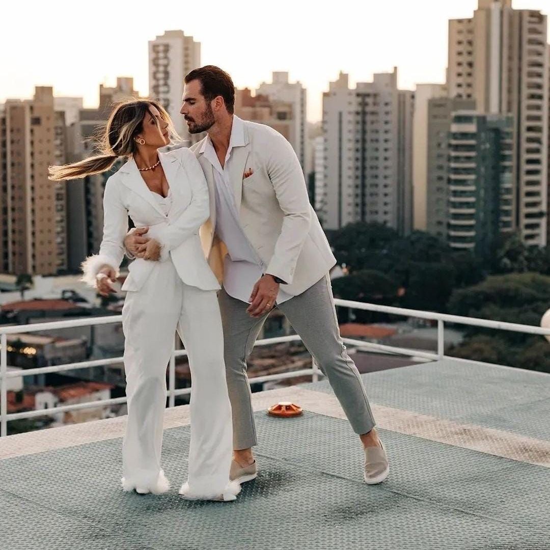 Casal vestido de branco em um terraço, demonstrando afeto com prédios da cidade ao fundo.