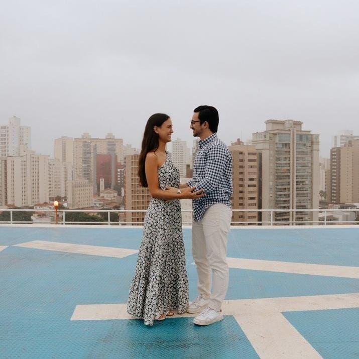 Casal de mãos dadas em um heliporto no telhado de um prédio, com arranha-céus da cidade ao fundo.