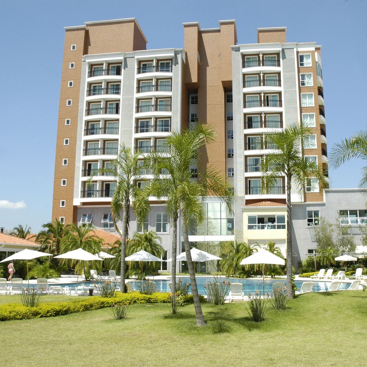 Hotel moderno com apartamentos, varandas, piscina, guarda-sóis brancos e palmeiras sob um céu límpido.