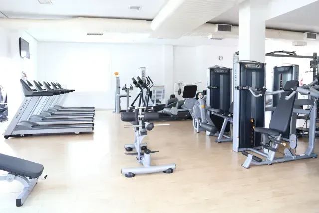 Interior de ginásio com esteiras, máquinas elípticas, máquinas de musculação e um banco sobre um piso de cor clara.