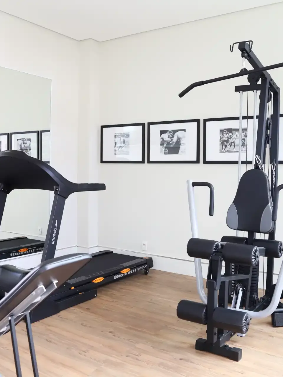 Academia em casa com esteira, aparelho de musculação e quadros na parede, em um ambiente bem iluminado.