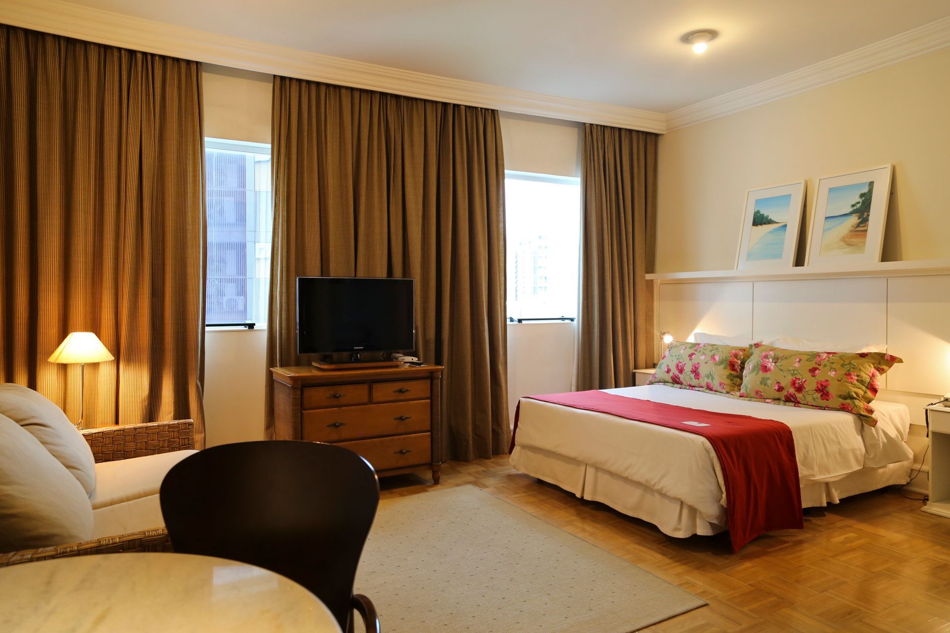 Quarto de hotel aconchegante com cama, TV, poltrona, abajur e cortinas nas janelas.