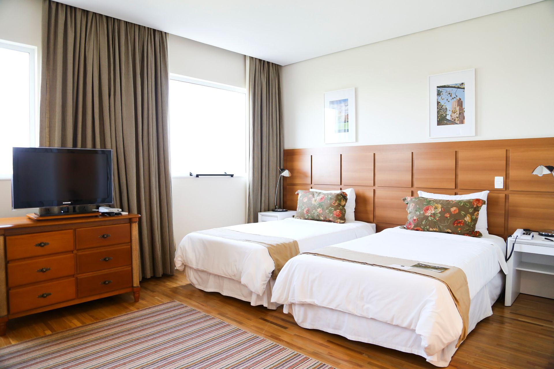 Quarto de hotel moderno com duas camas de solteiro, cabeceiras de madeira, cômoda, TV e janela com cortinas.
