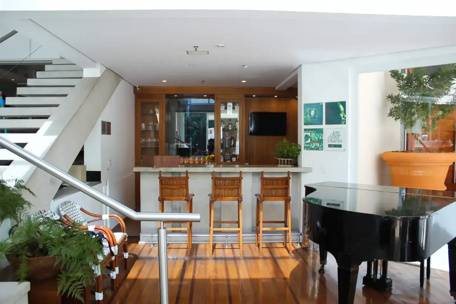 Interior com bar, piano, escadaria e plantas em vasos. Piso de madeira e banquetas de bar.