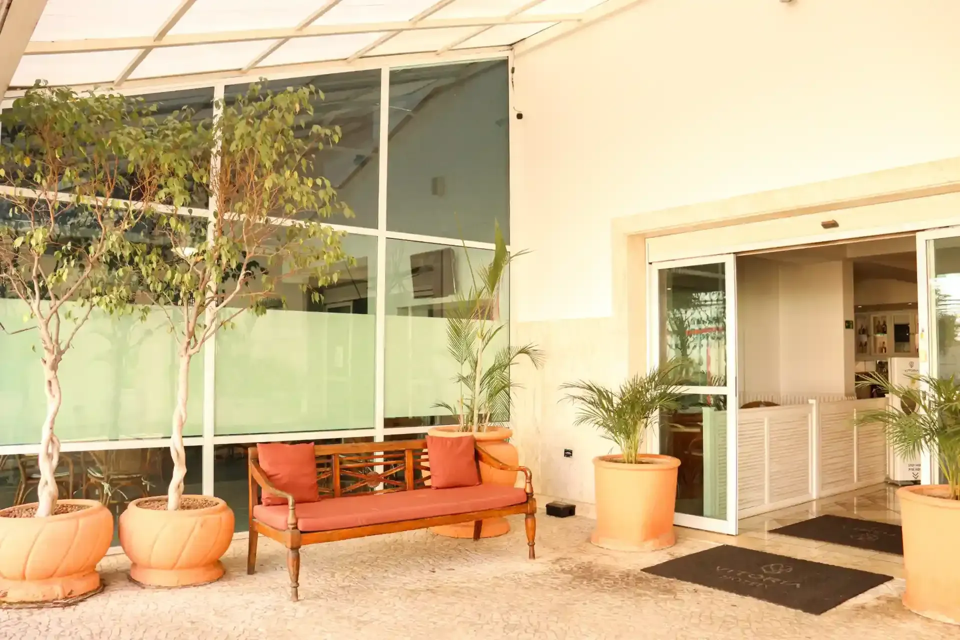 Entrada externa do edifício com árvores em vasos, um banco vermelho e portas de vidro sob um toldo.