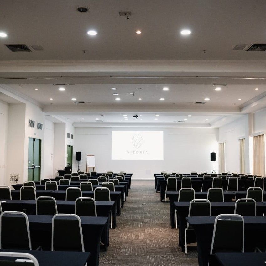Sala de conferências vazia com fileiras de cadeiras pretas voltadas para uma tela de projeção.
