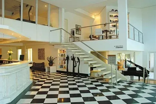 Piso quadriculado em preto e branco em um saguão moderno. Uma escada leva ao segundo andar. Um piano de cauda está localizado nas proximidades.
