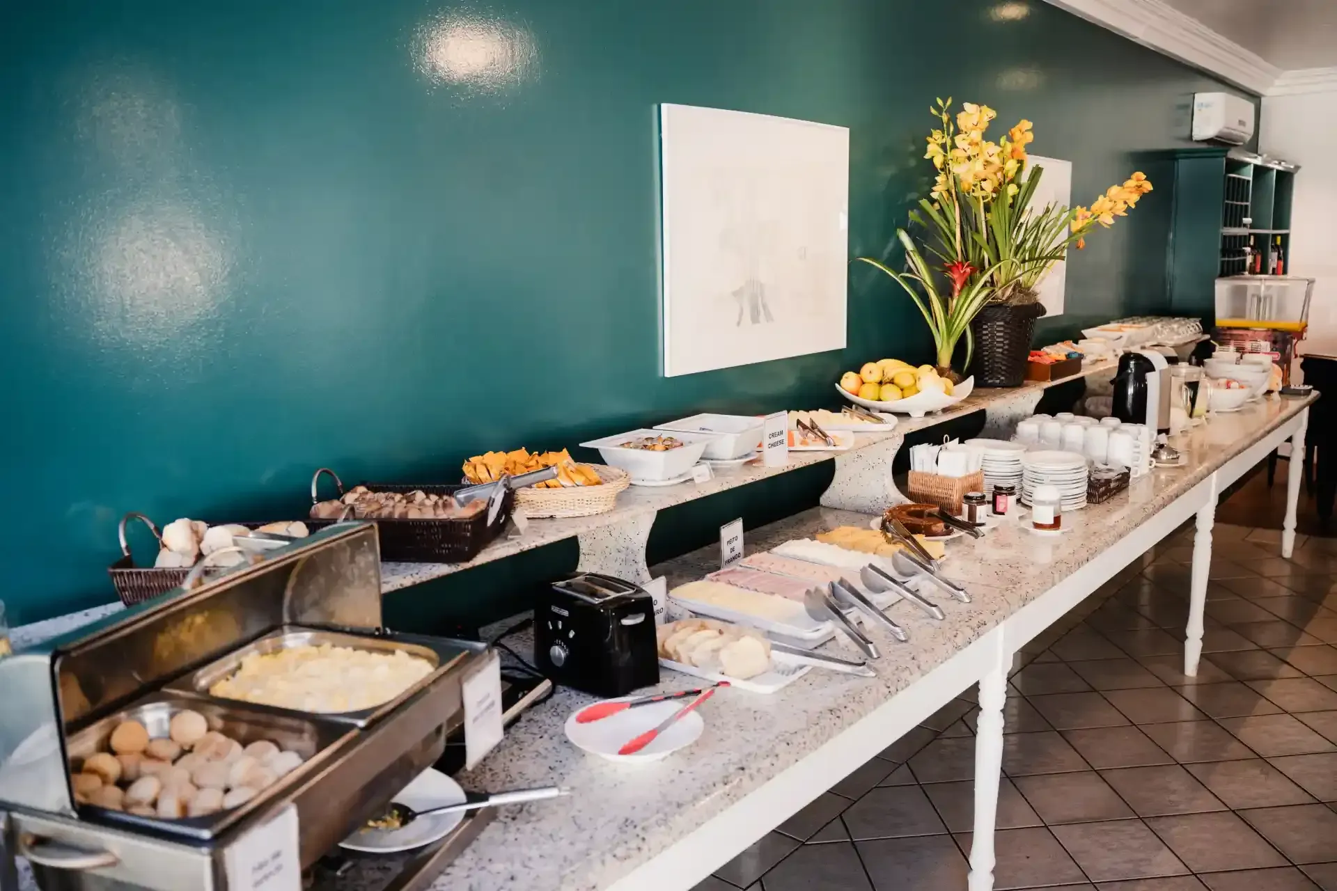 Buffet de café da manhã com doces, frutas, café e flores em uma longa mesa em uma sala de jantar com paredes verde-azuladas.