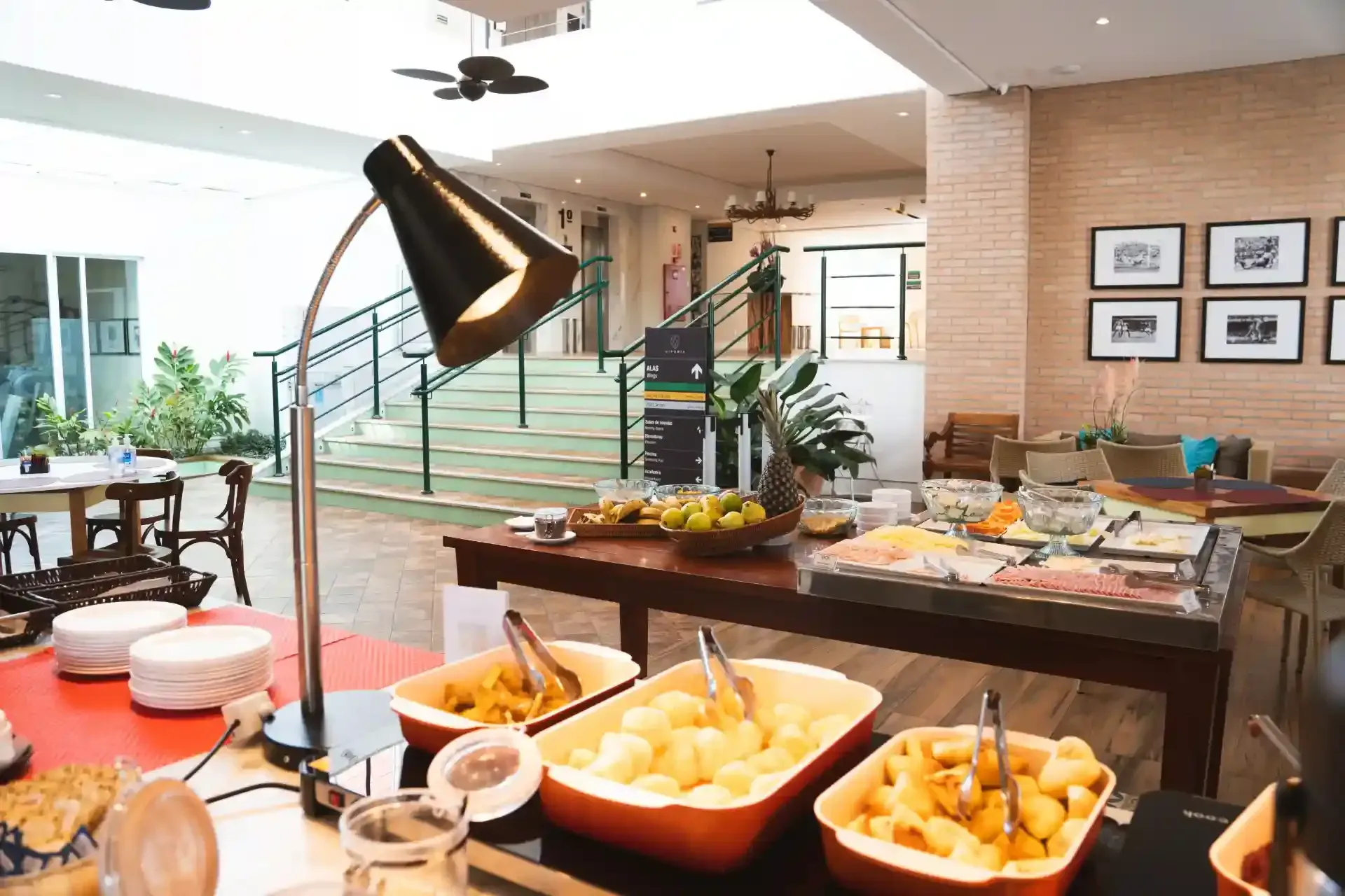 Buffet de café da manhã com diversos itens alimentares sobre uma mesa de madeira, sob uma lâmpada preta.