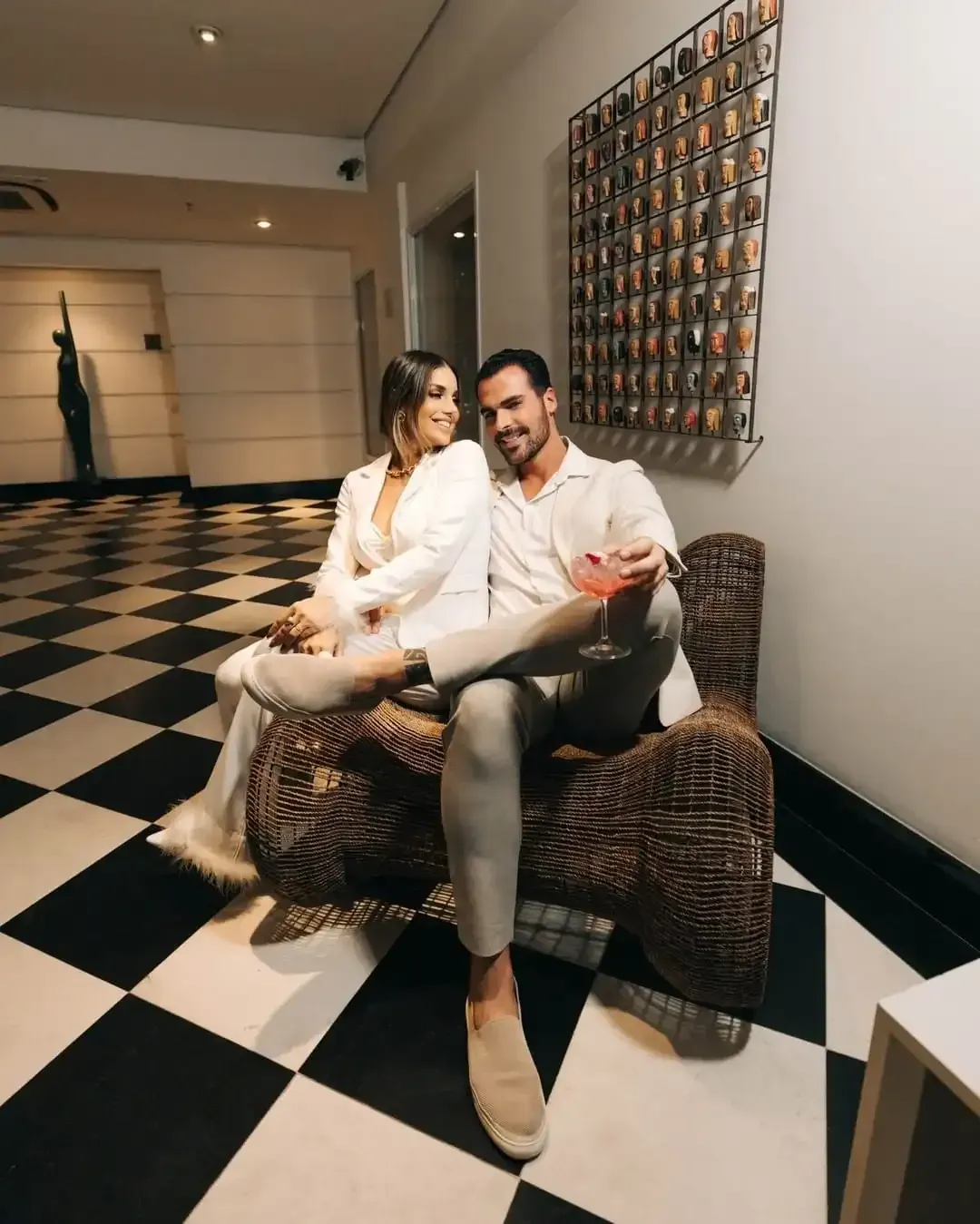 Casal com roupas em tons neutros sentado em uma cadeira estampada em um saguão com piso quadriculado em preto e branco, olhando um para o outro.