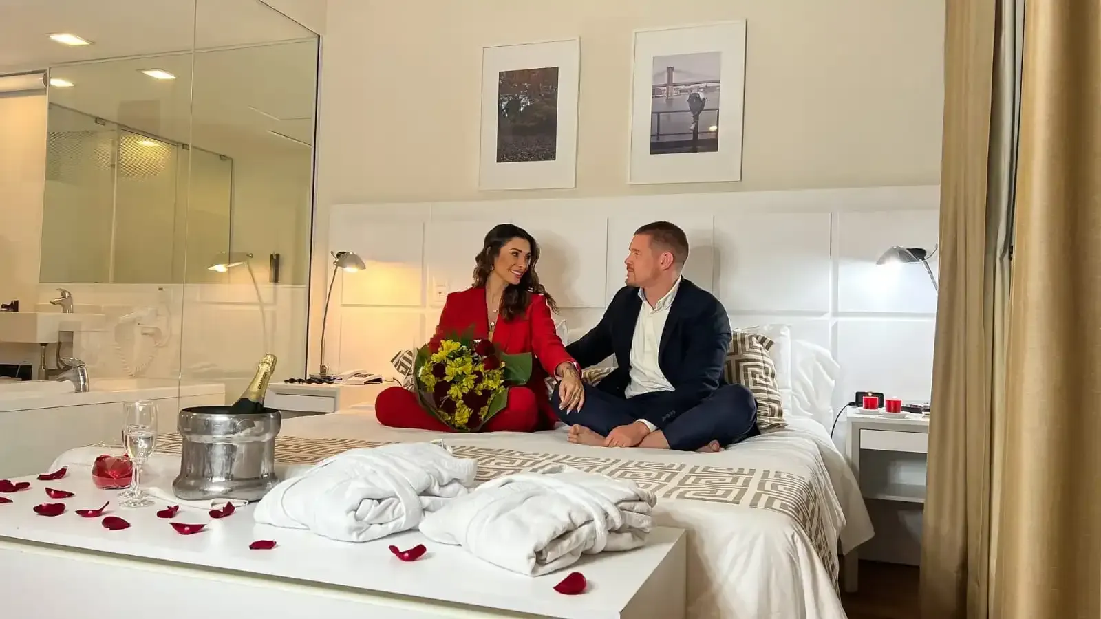 Um casal está sentado na cama, abraçado. Champanhe, flores e pétalas de rosa decoram o quarto.