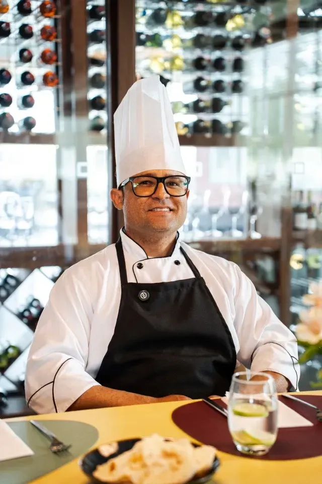 Chef de cozinha com chapéu branco e avental preto, sentado à mesa em um restaurante, sorrindo.