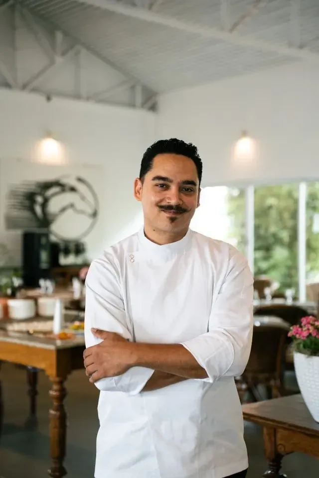 Homem de jaleco branco sorrindo com os braços cruzados na cozinha iluminada de um restaurante.