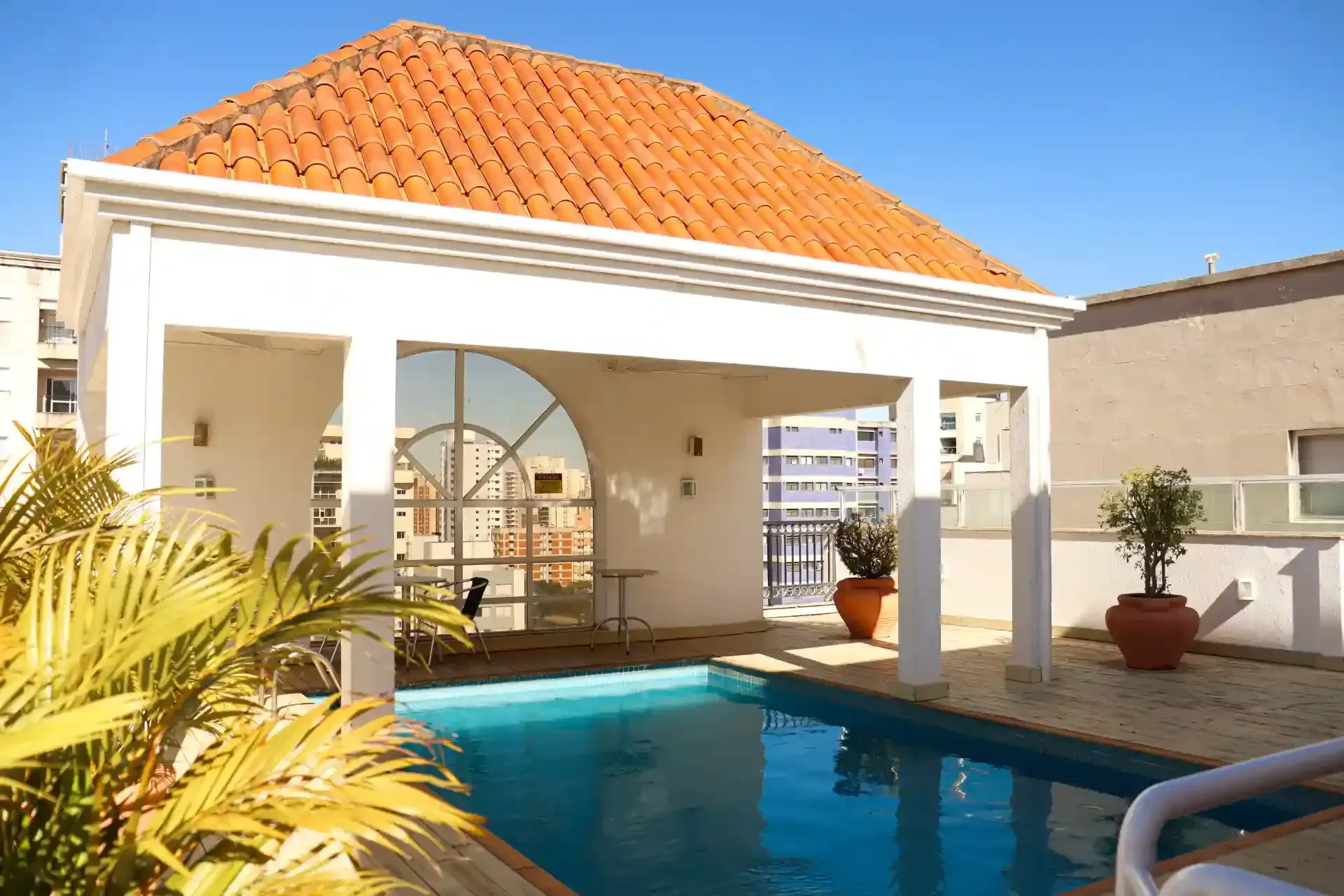 Pátio moderno com gazebo de telhas vermelhas ao lado de uma piscina azul sob a luz do sol.