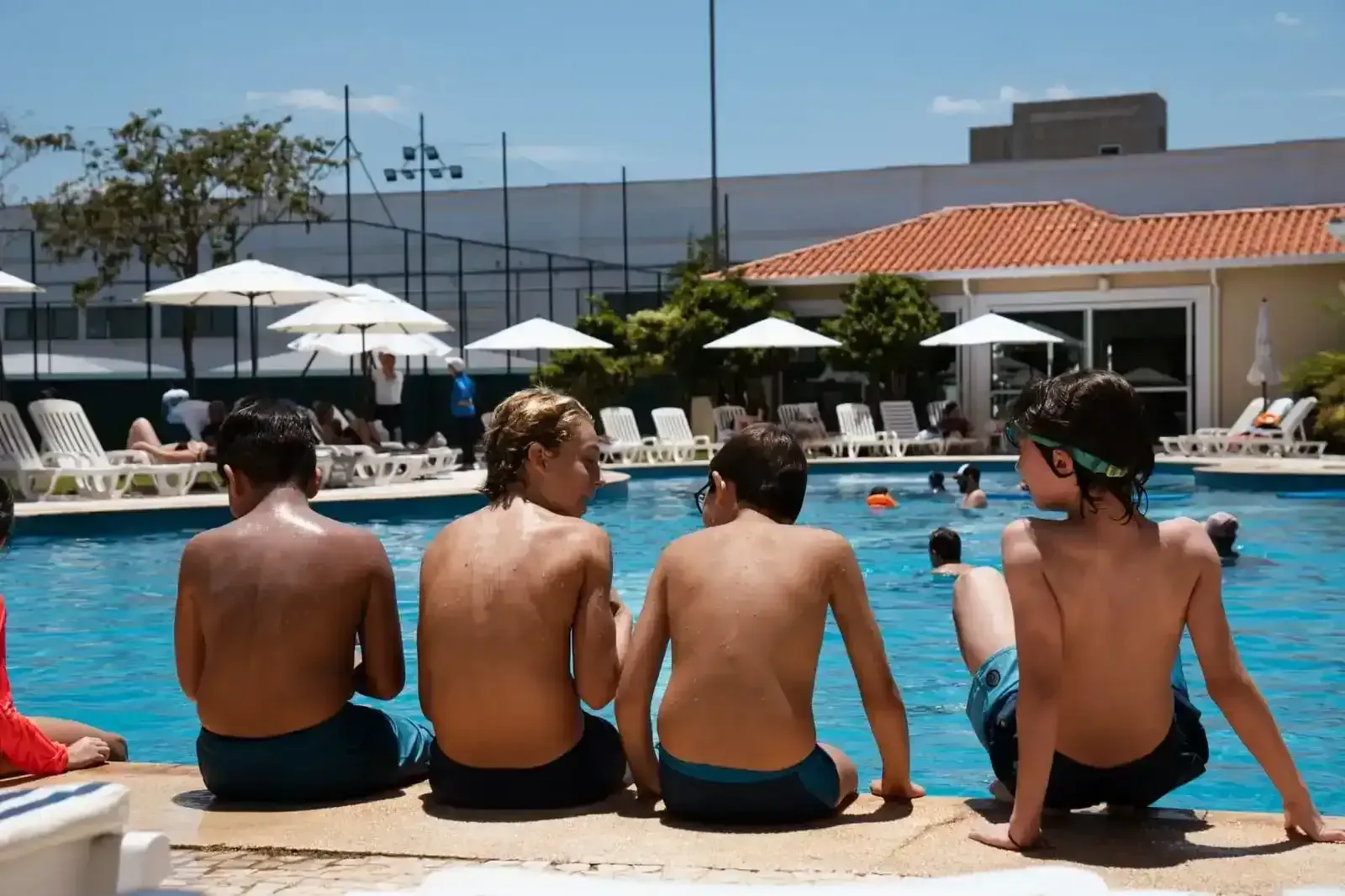 Quatro pessoas sem camisa sentam-se à beira da piscina, com água azul e guarda-sóis ao fundo.