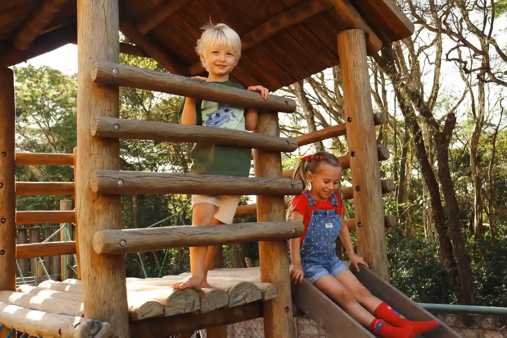 Duas crianças brincando em um parquinho de madeira: o menino em pé, a menina escorregando. Cores verde, azul e vermelha visíveis. Floresta ao fundo.