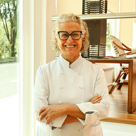 Um chef sorridente, de jaleco branco, está de braços cruzados na cozinha iluminada de um restaurante.