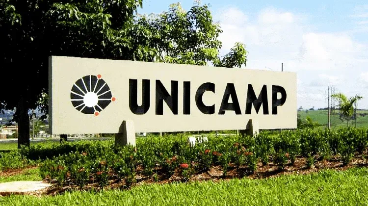 Placa com a palavra UNICAMP e o logotipo em frente a um gramado verde com árvores.