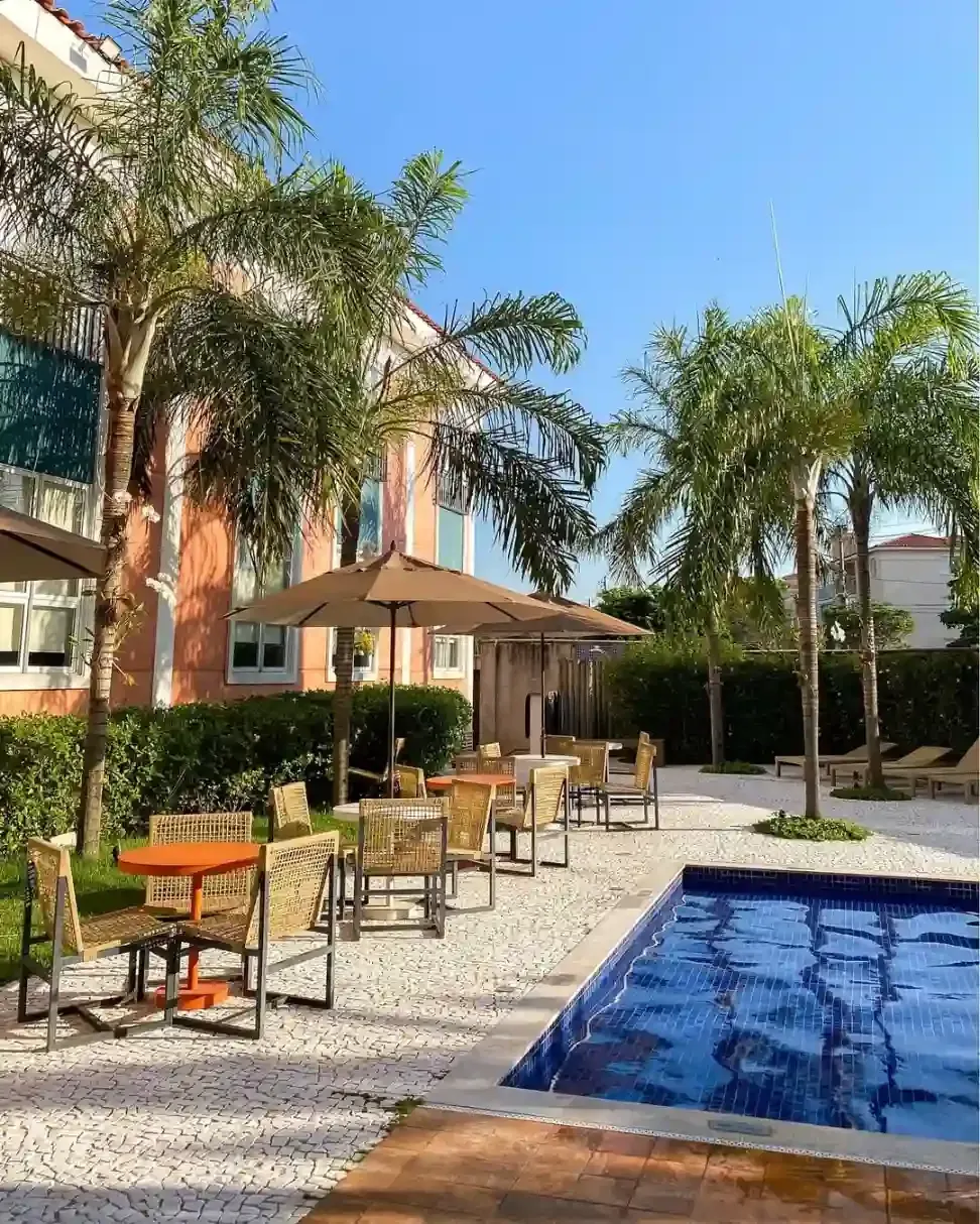 Pátio à beira da piscina com palmeiras, espreguiçadeiras e guarda-sóis cor de areia ao lado de uma piscina azul.
