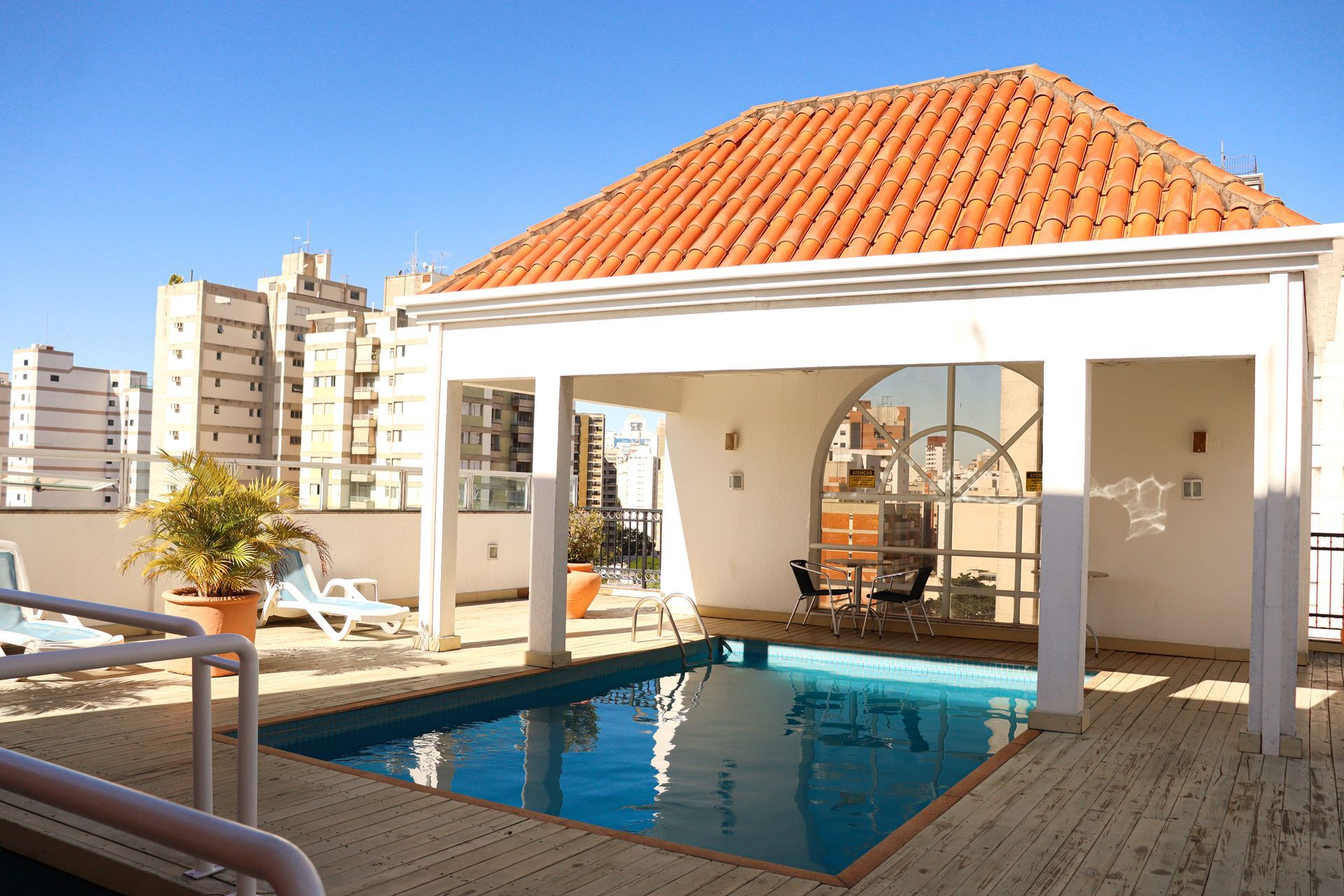 Vista da piscina retangular, rodeada por plantas e edifícios, sob um céu azul.