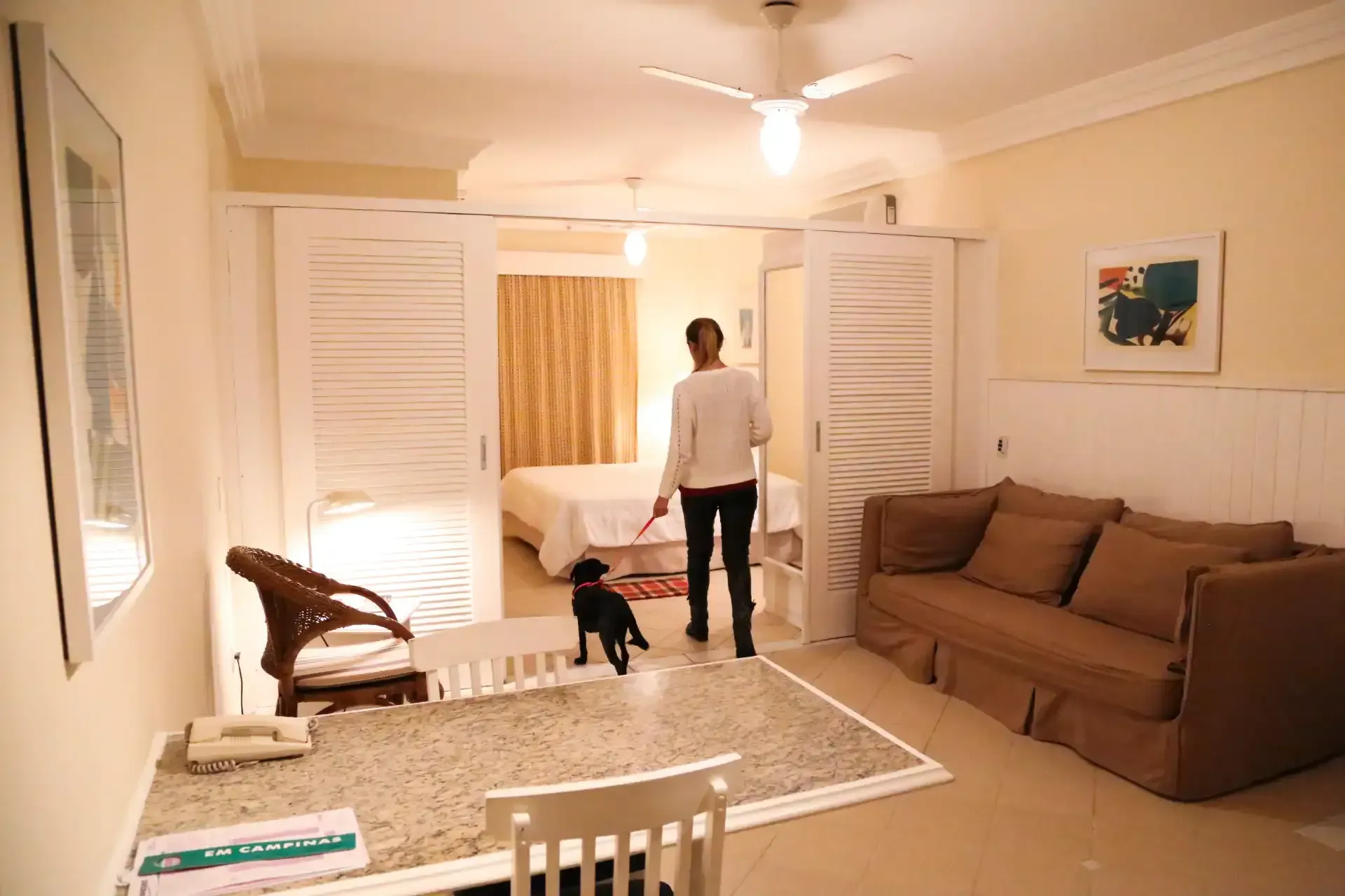 Sala de estar com um sofá marrom, mesa de jantar e uma pessoa em pé perto de uma criança junto à porta.