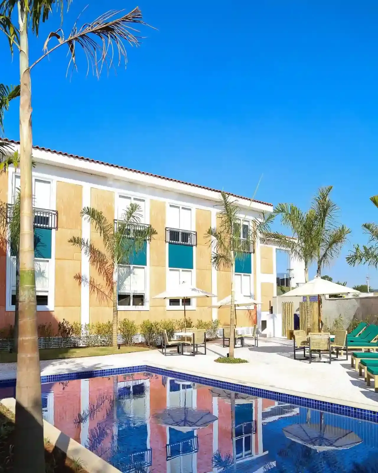 Vista da piscina de um prédio amarelo de um resort com palmeiras sob um céu azul brilhante.