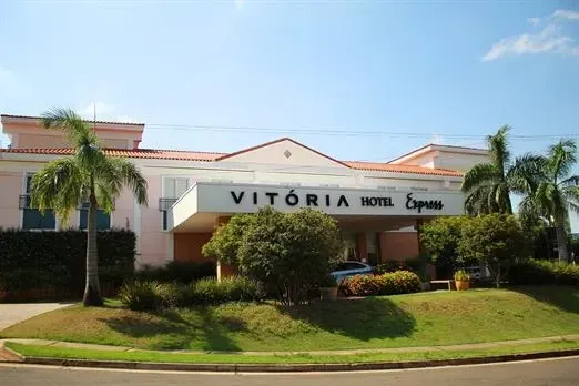 Entrada principal do Hotel Vitória, um prédio baixo e branco com palmeiras e um gramado verde.