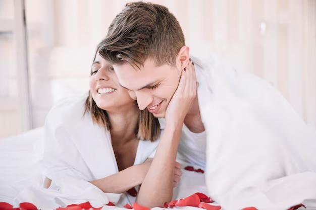 Casal na cama, sorrindo e se abraçando sob lençóis brancos.