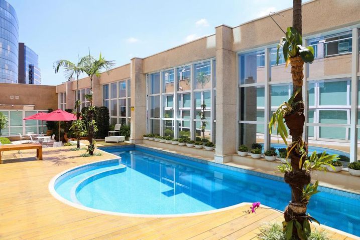 Área da piscina e pátio com edificação. Piscina azul, deck bege, janelas e palmeiras.