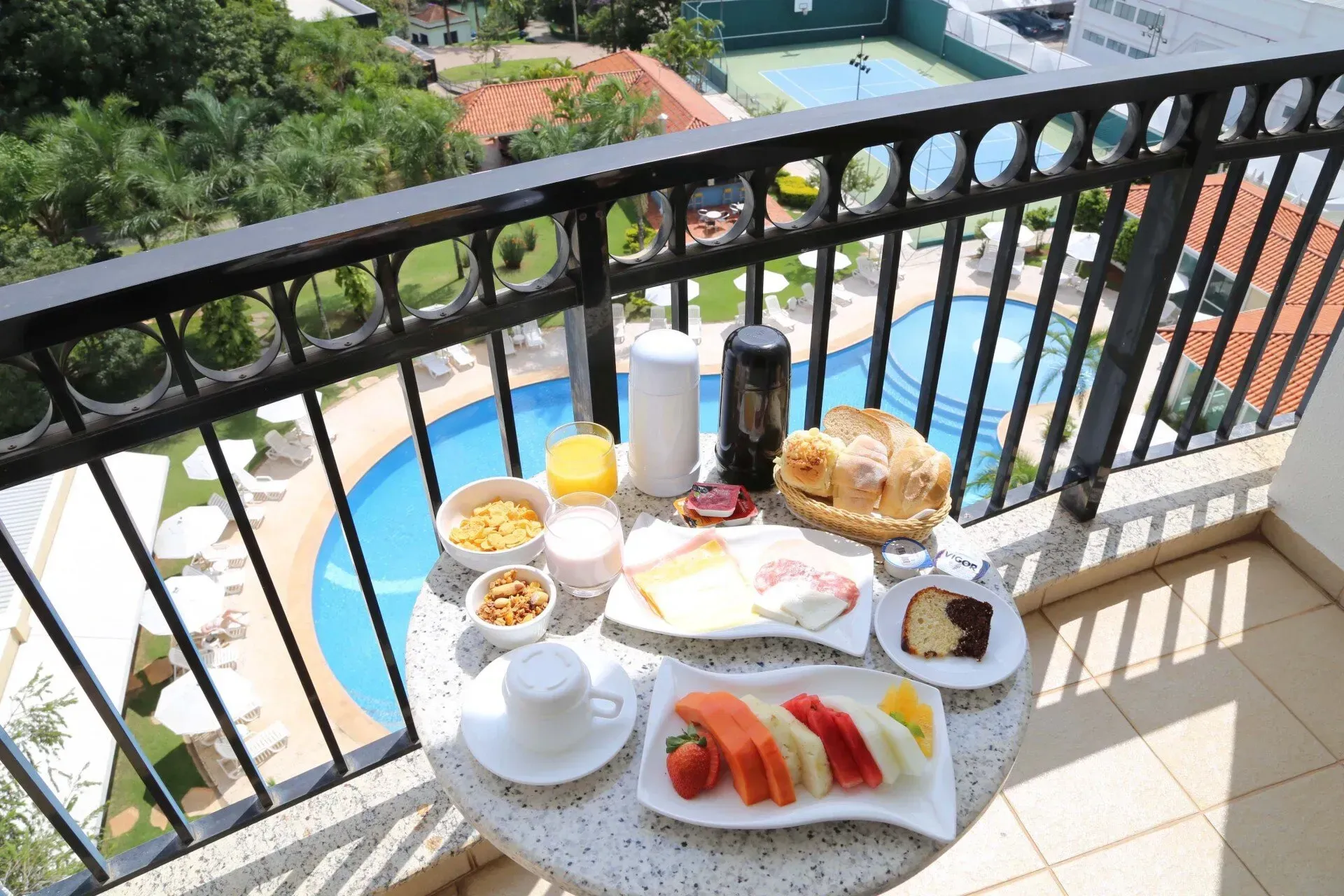 Bandeja de café da manhã na varanda com croissants, frutas, café, suco e vista para a piscina.