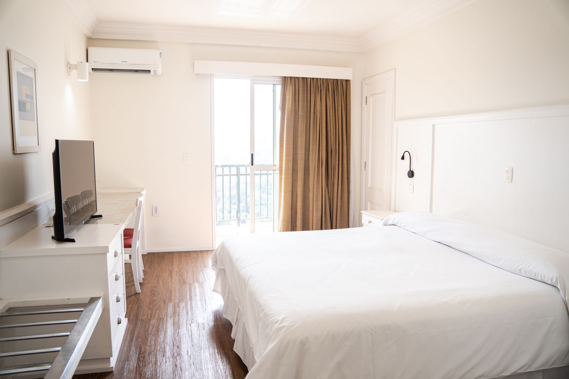 Quarto de hotel luminoso com cama branca, escrivaninha, TV e portas de varanda à luz do dia.