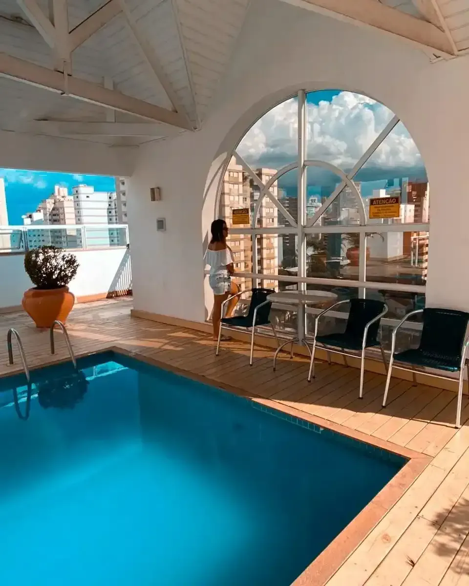 Piscina coberta ao lado de uma janela em arco com vista para a cidade, espreguiçadeiras brancas e plantas em vasos.