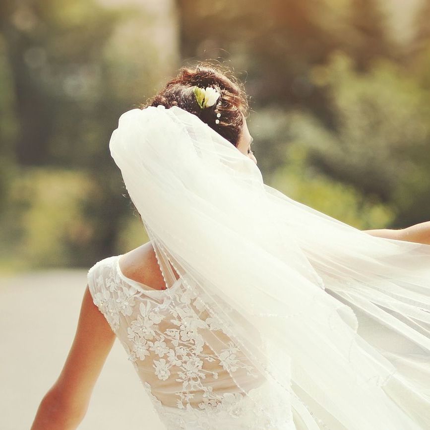 Noiva em vestido de renda e véu longo, em pé ao ar livre sob a luz suave do sol.