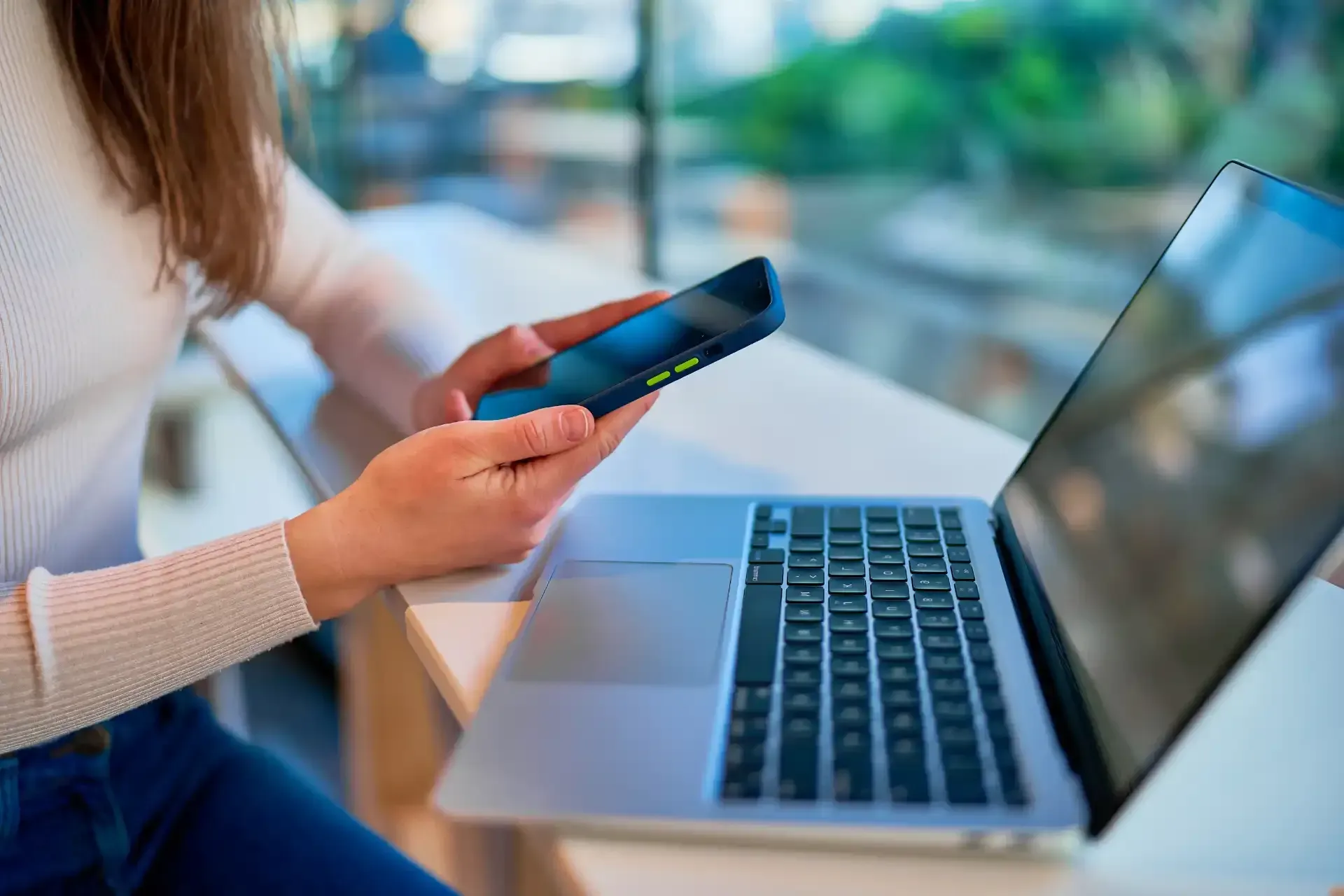 Mulher segurando o celular e usando um laptop em uma mesa perto da janela.
