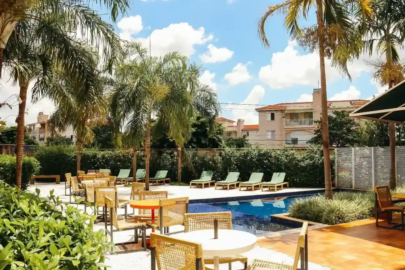 Piscina ensolarada de resort com palmeiras, espreguiçadeiras e área de estar no terraço à beira de uma piscina azul.