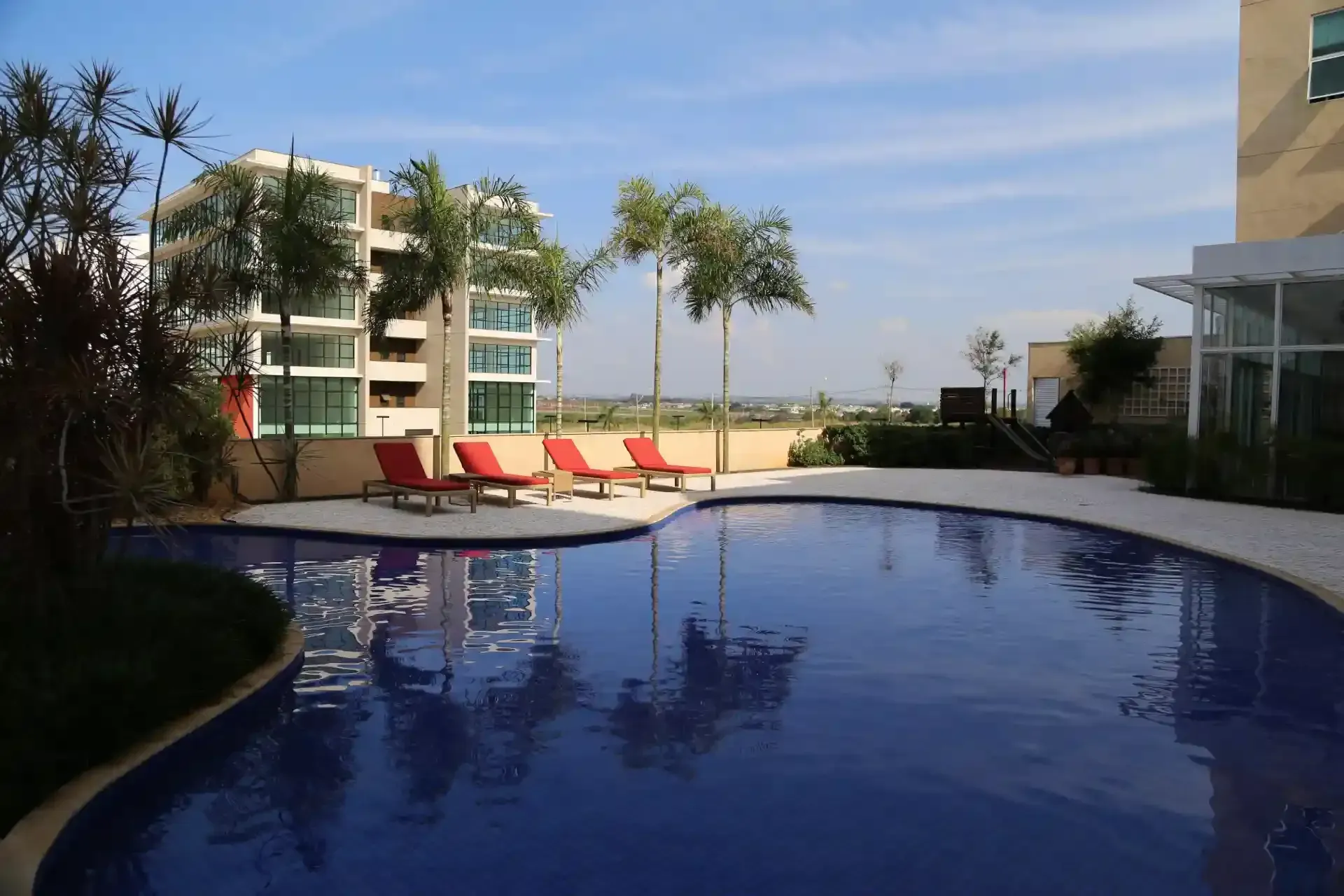 Piscina com espreguiçadeiras, construção e palmeiras sob um céu azul.