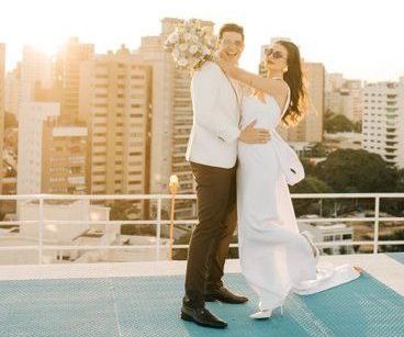 Casal vestido de branco posando em um terraço ao pôr do sol, com prédios da cidade ao fundo.