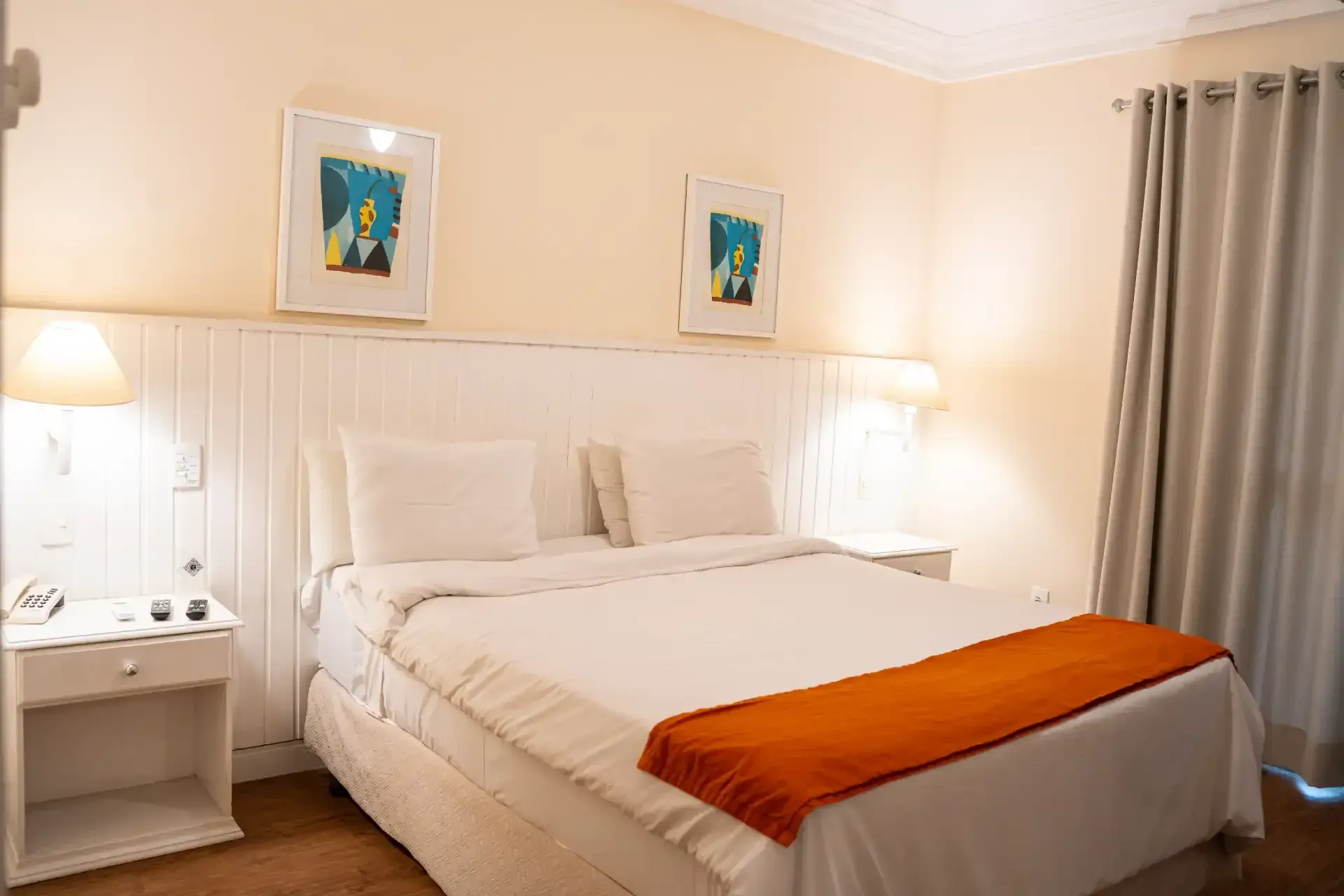 Cama de hotel impecavelmente arrumada com lençóis brancos e uma manta laranja em um quarto bege claro.