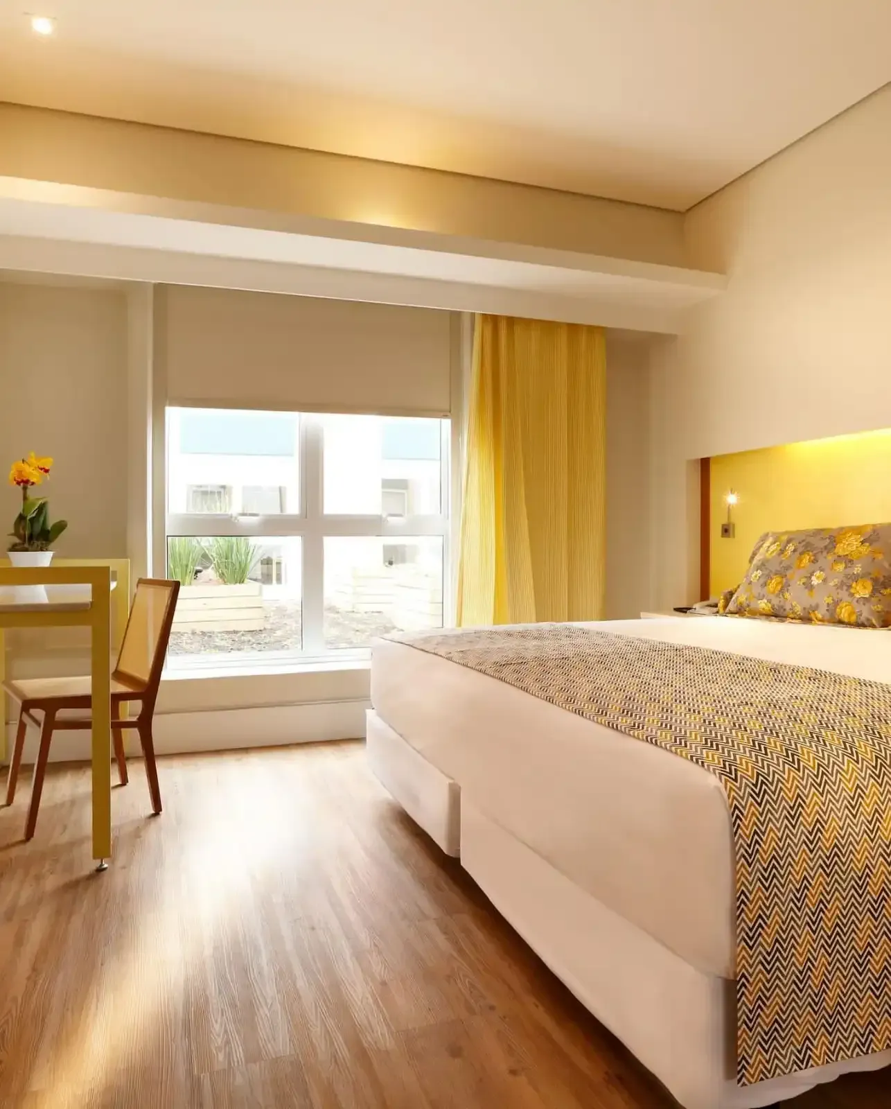 Quarto moderno e luminoso com uma cama grande, recanto com janela, escrivaninha e piso de madeira em tons quentes.