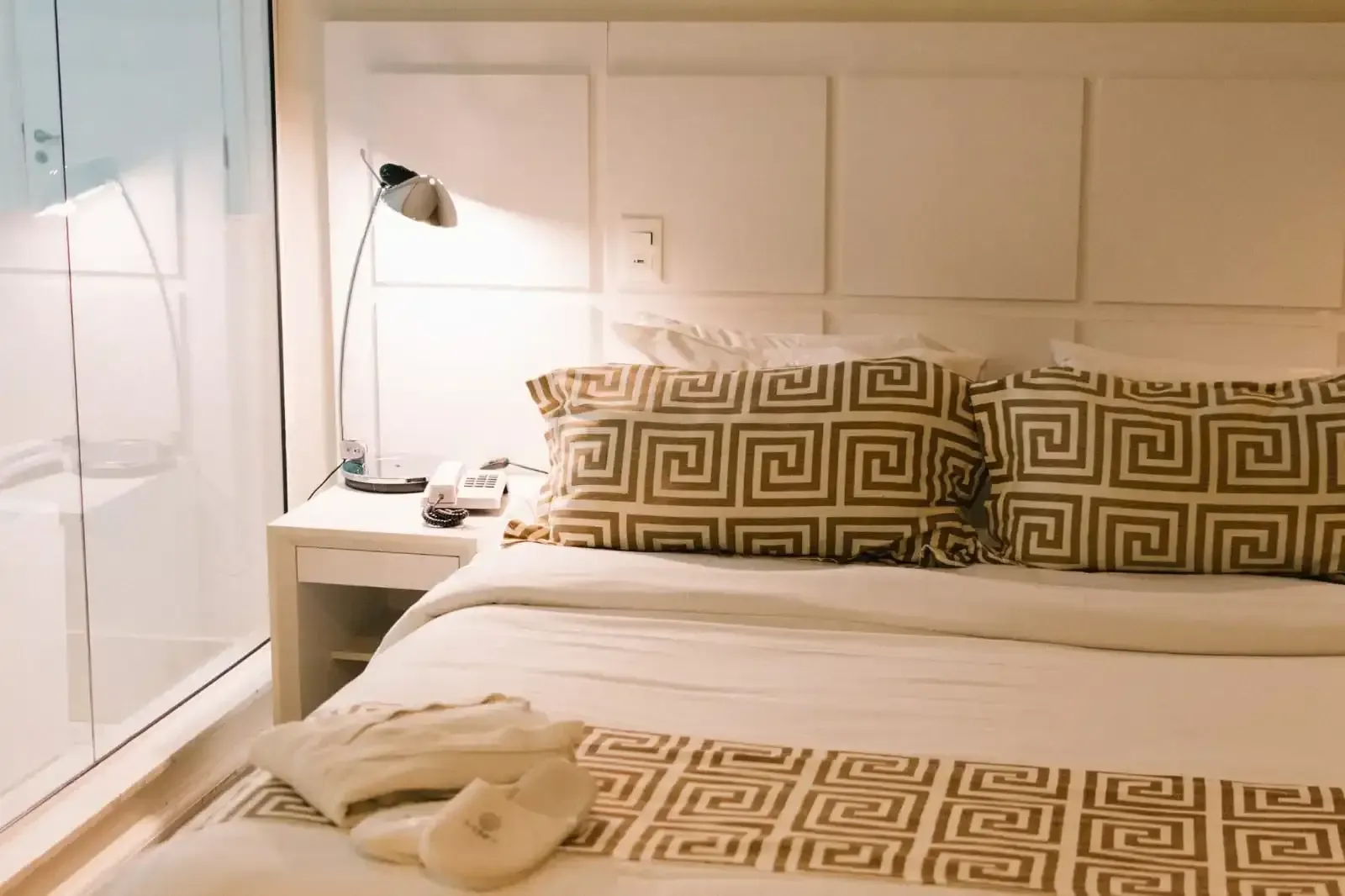 Quarto com cabeceira branca, cama com almofadas e manta estampadas. Mesa de cabeceira com abajur e telefone.
