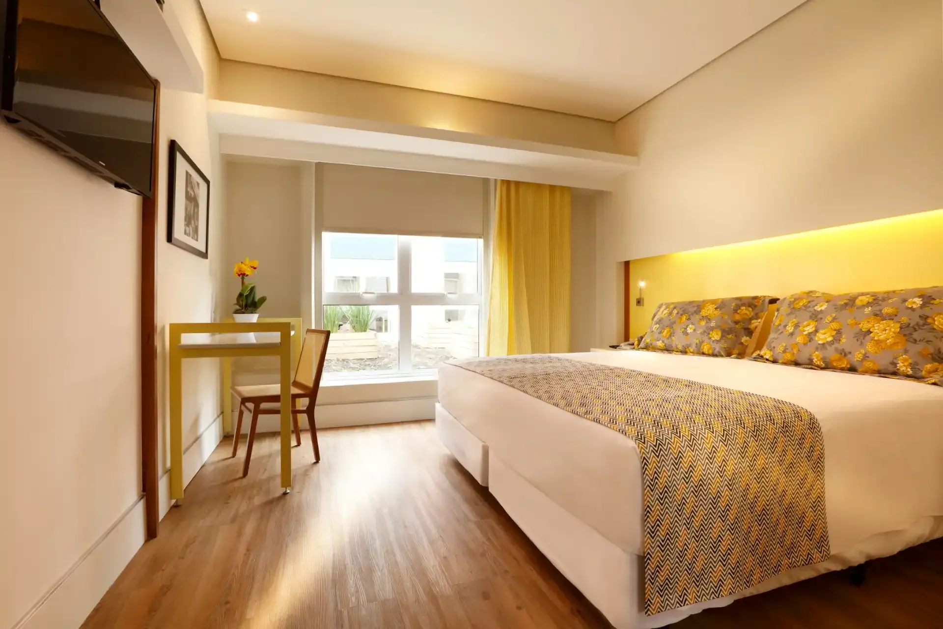 Quarto de hotel com cama, escrivaninha e janela com cortinas amarelas.