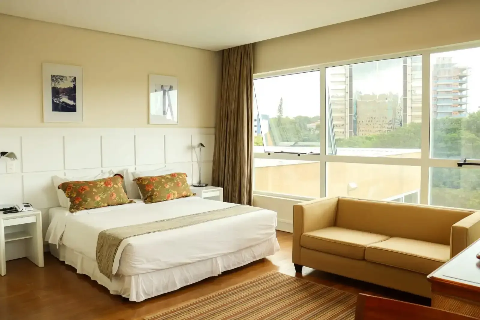 Quarto de hotel com cama, sofá pequeno e janelas amplas com vista para a cidade.