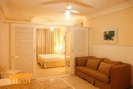 Sala de estar aconchegante com sofá bege, mesinha de centro, quadros e um quarto adjacente com persianas e cortinas brancas.