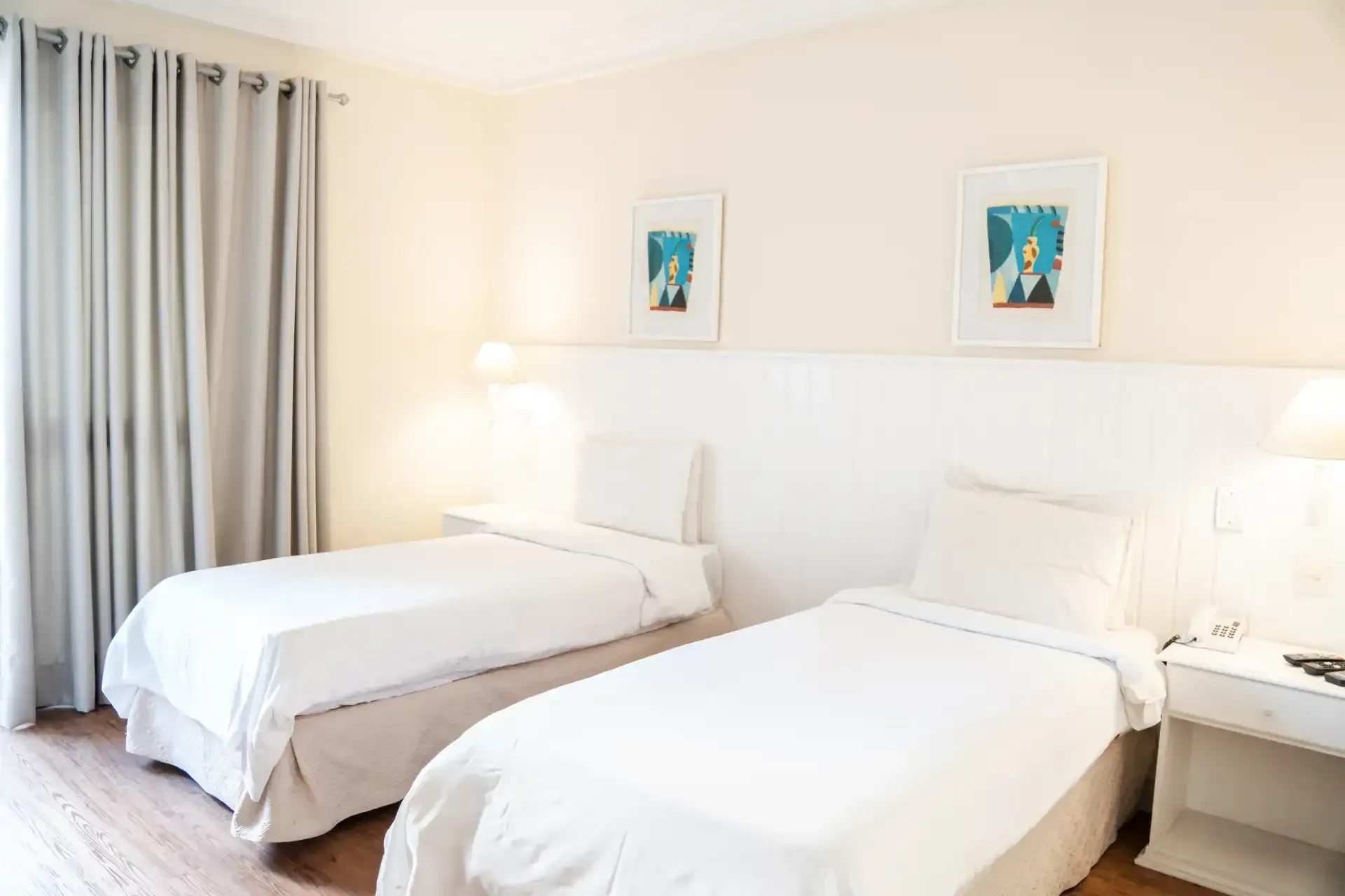 Quarto claro e branco com duas camas de solteiro, abajur e obras de arte emolduradas na parede.