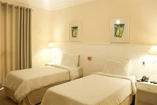 Quarto de hotel com duas camas de solteiro, paredes em tons creme, abajures, obras de arte emolduradas e cortinas fechadas.