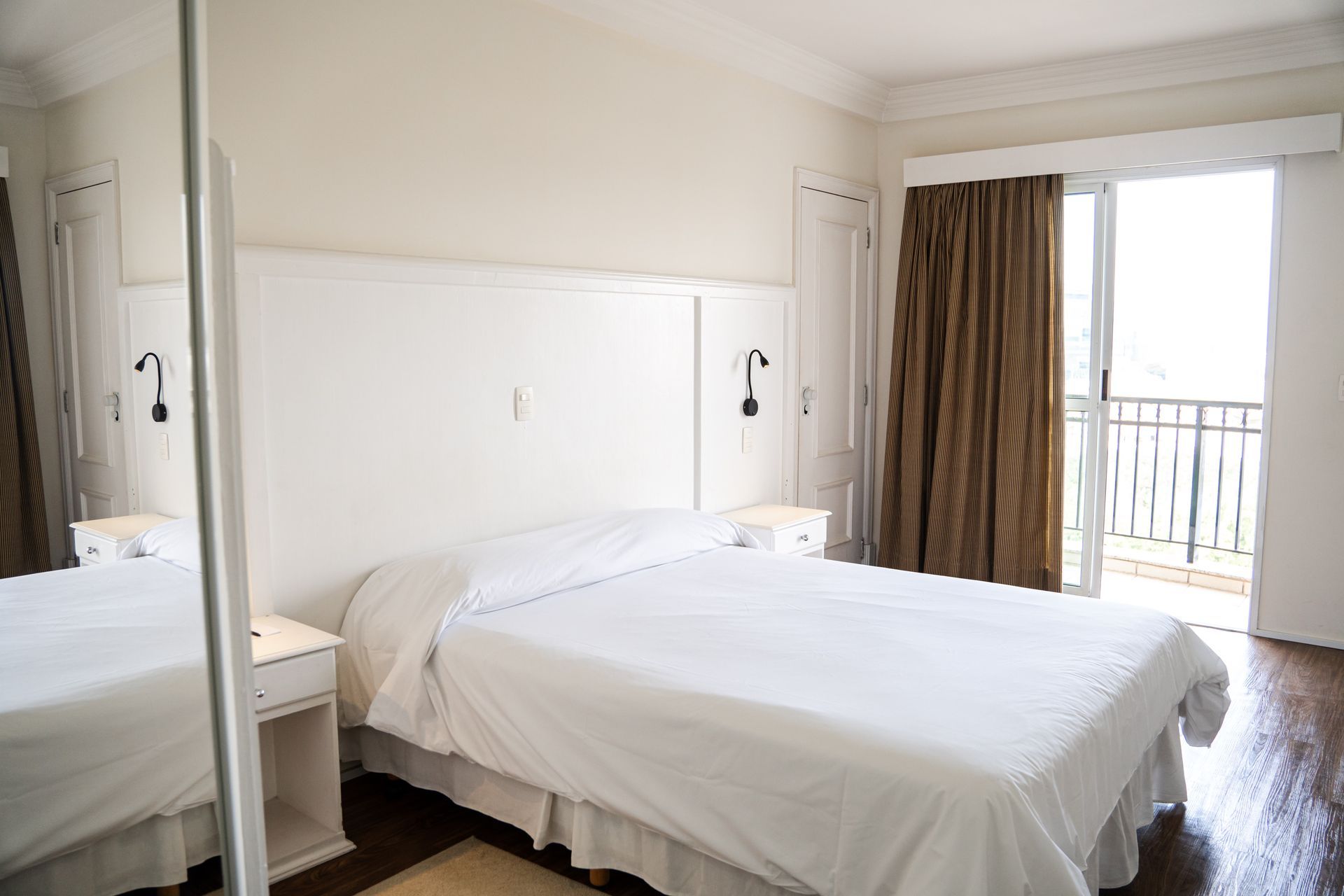 Quarto luminoso com uma cama branca impecavelmente arrumada, guarda-roupa com espelho e porta para varanda com cortinas.