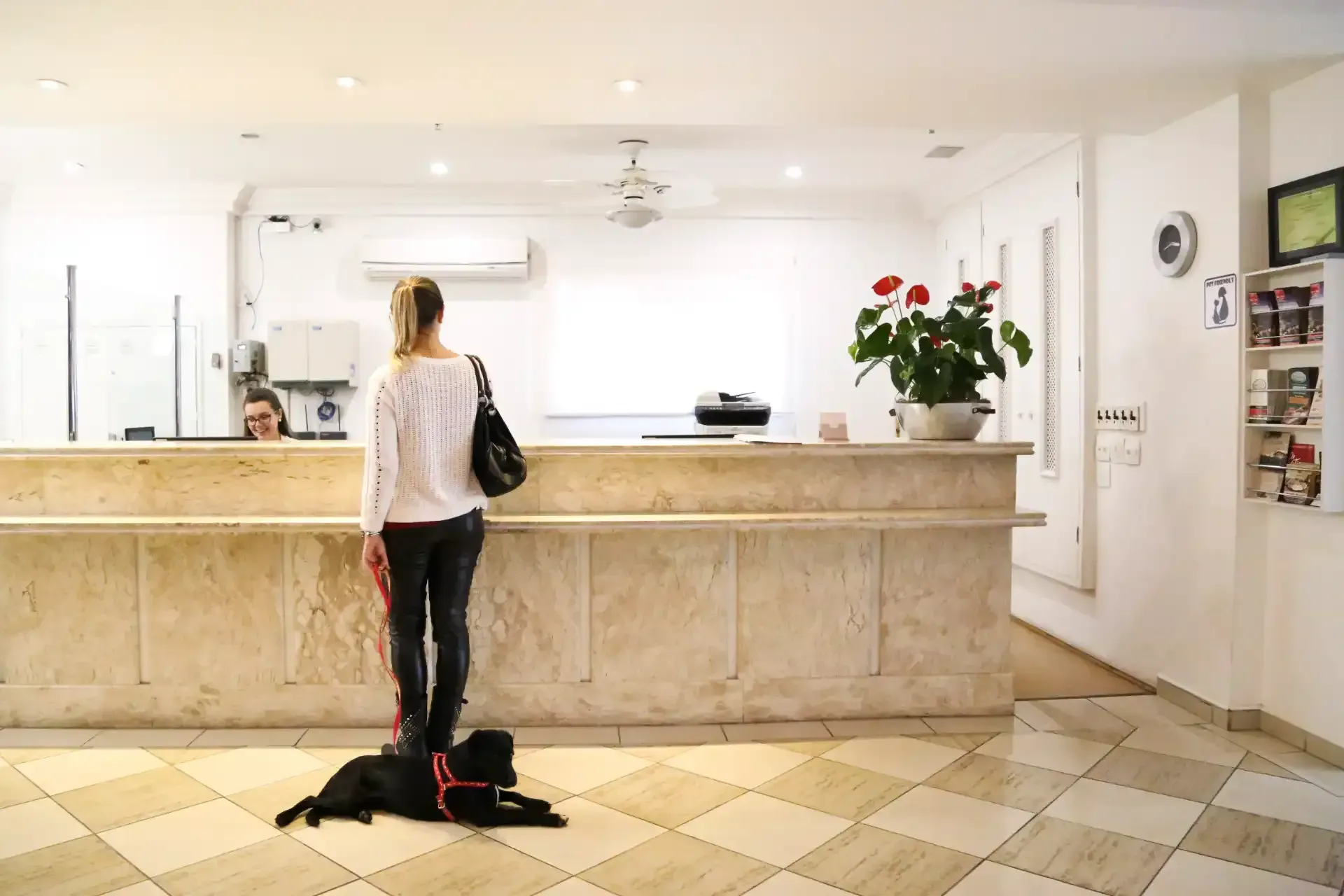 Pessoa em pé na recepção de um hotel com uma bolsa preta no chão e flores vermelhas no balcão.