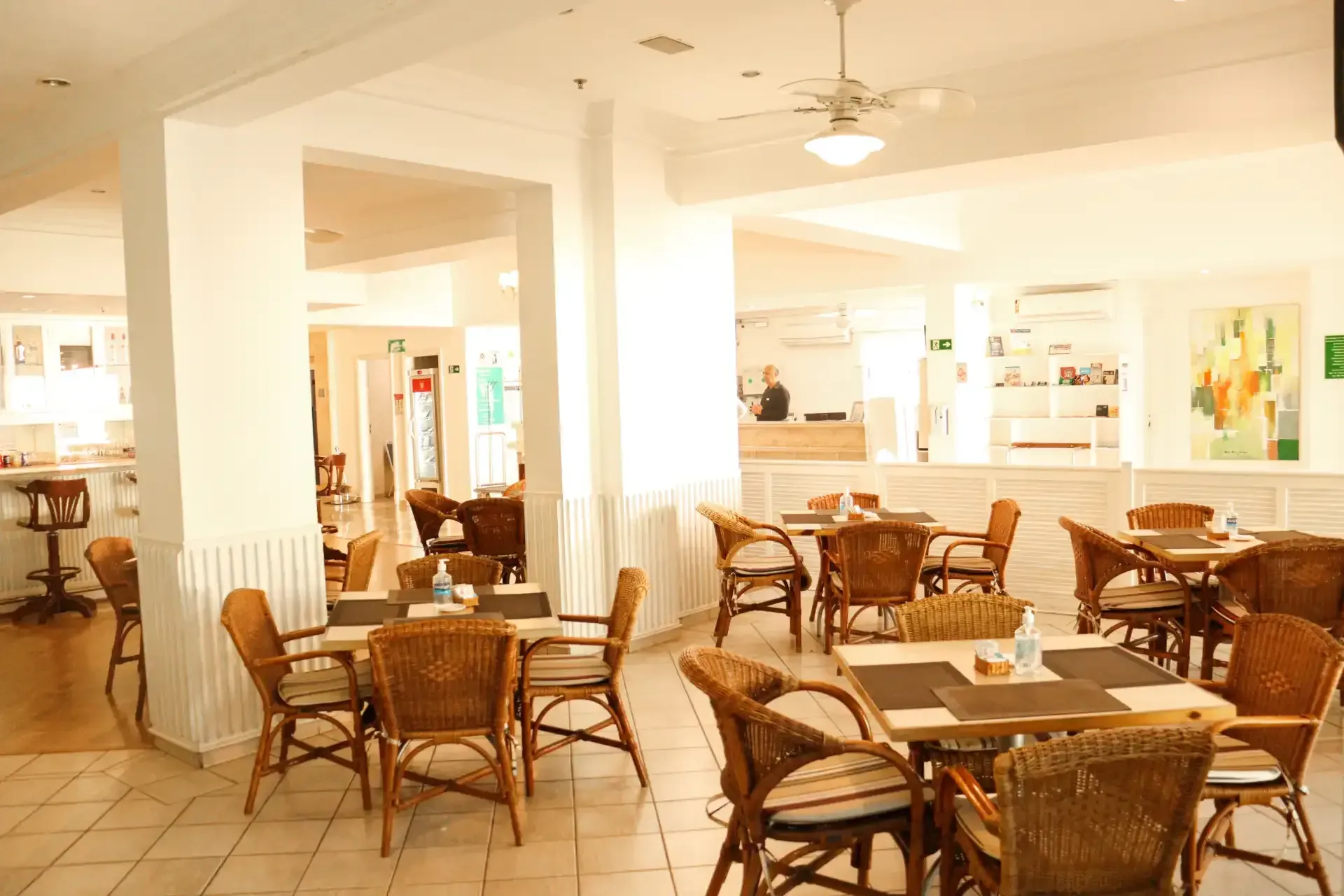 Restaurante luminoso e arejado com cadeiras de vime, mesas de madeira e colunas brancas em um salão de jantar banhado de sol.
