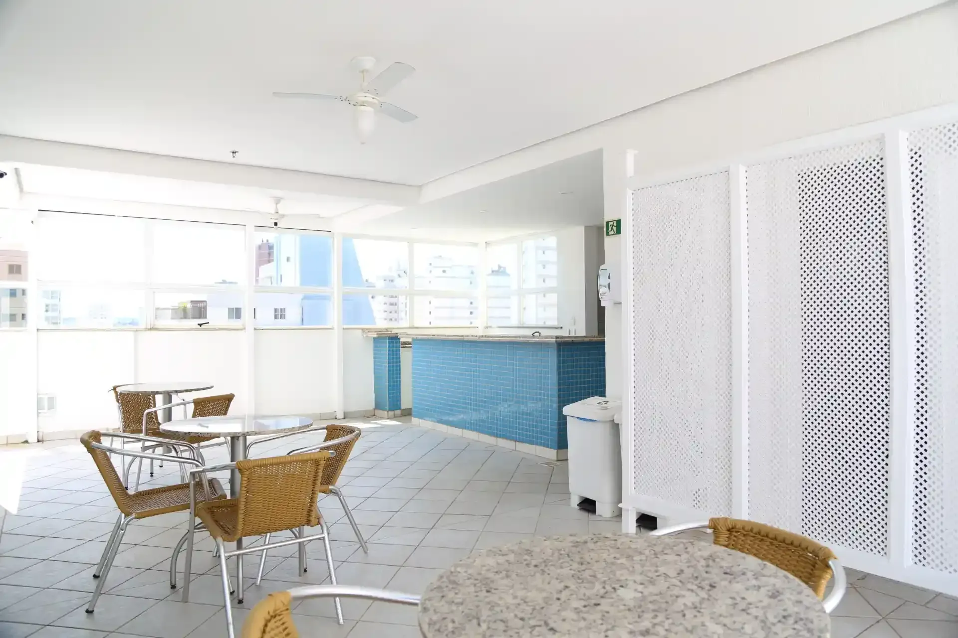 Interior de café luminoso e branco com cadeiras de vime, mesas redondas, piso de azulejo e balcão de bar azul.
