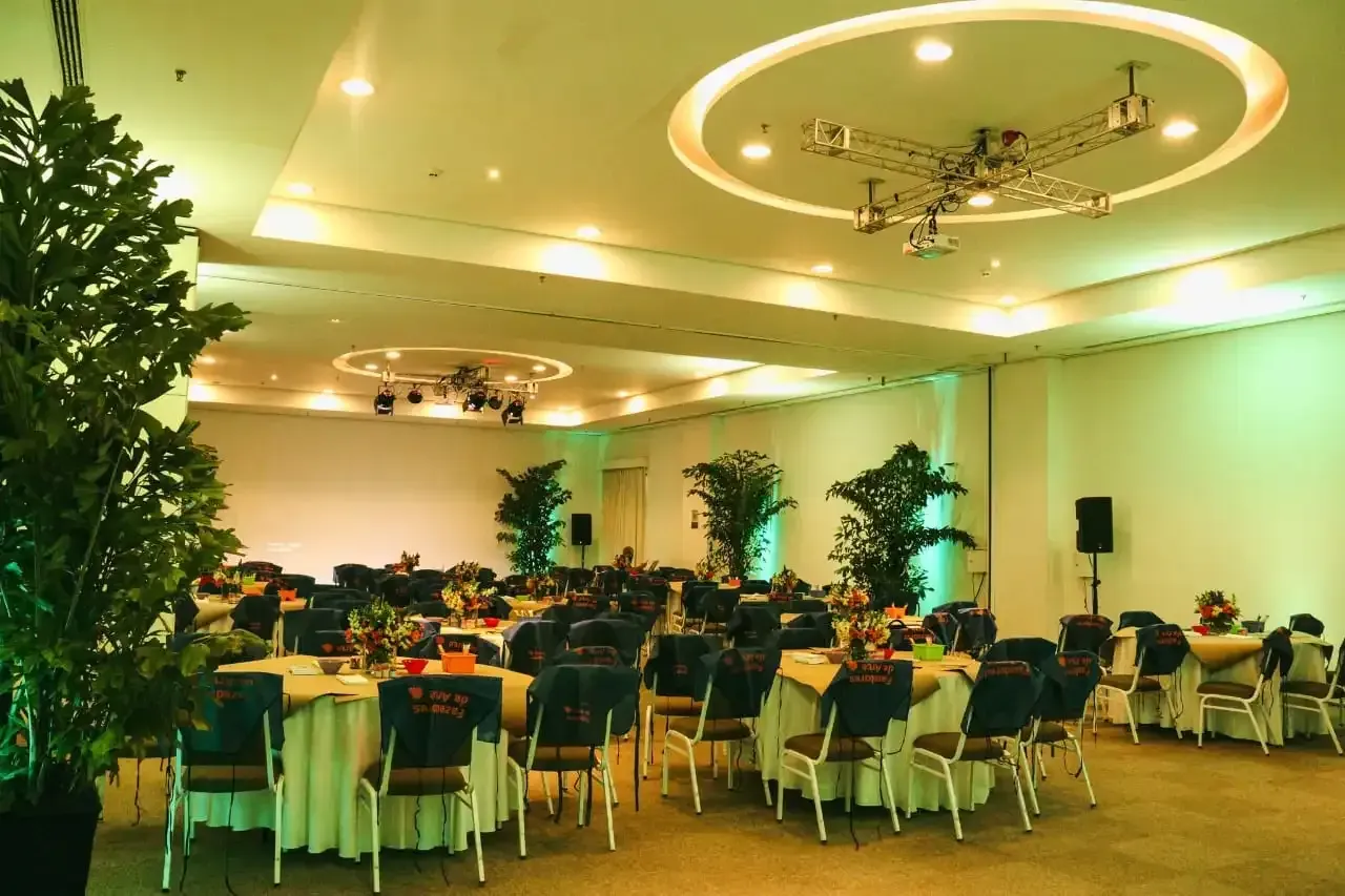 Salão de banquetes elegante com mesas redondas, cadeiras brancas, iluminação aconchegante no teto e detalhes em verde.